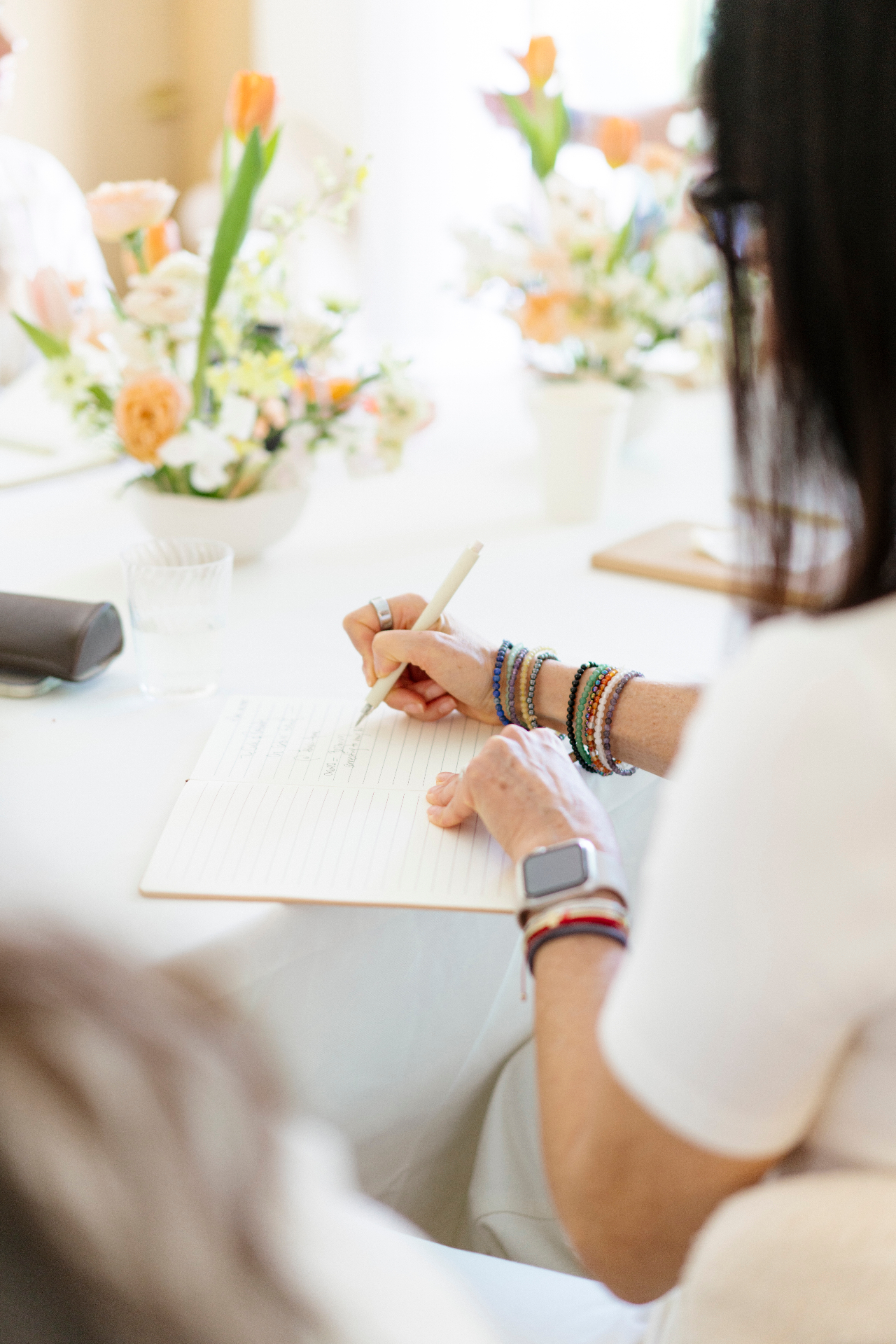A woman writing in an open notebook at a sunlit table set with tulips and soft florals.